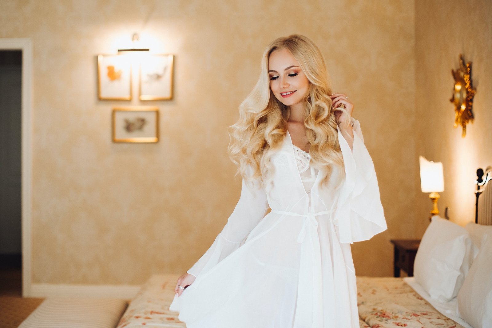 Model wearing a white lace bridal-style set in a warmly lit bedroom.
